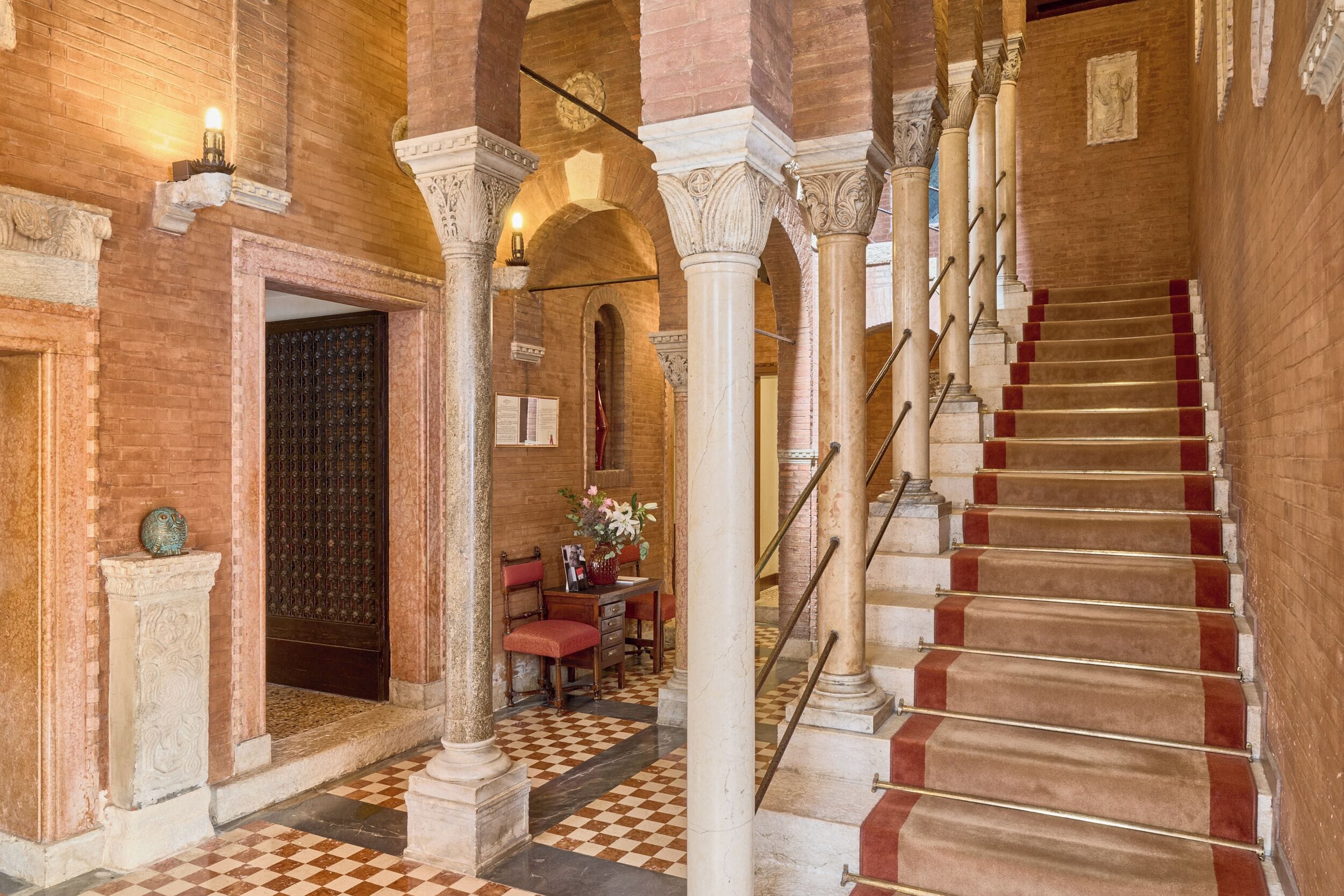 Una gran escala amb passades revestides de catifes vermelles, flanquejada per columnes i arcs blancs, que condueix a una sala amb un paviment de pissos quadriculats i un escriptori amb un ram de flors.