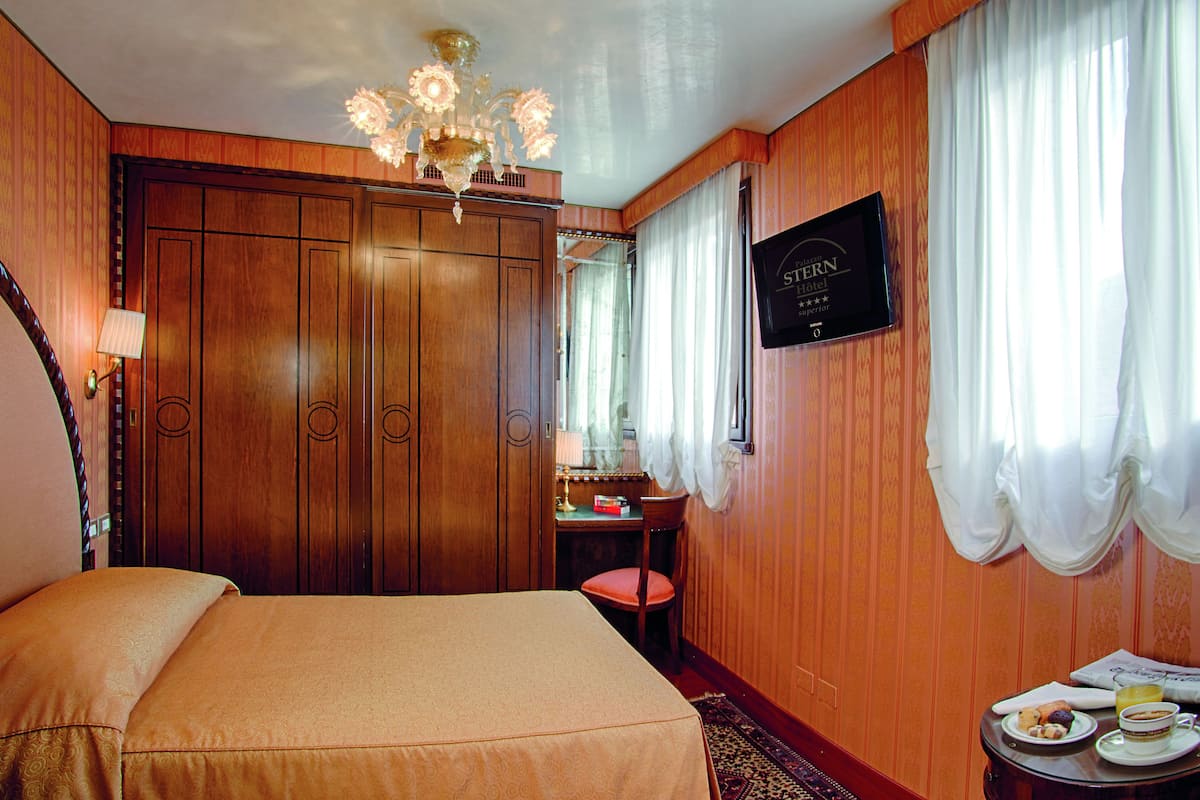 A hotel room with a bed, a desk, a chair, a chandelier, a sign, and curtains.