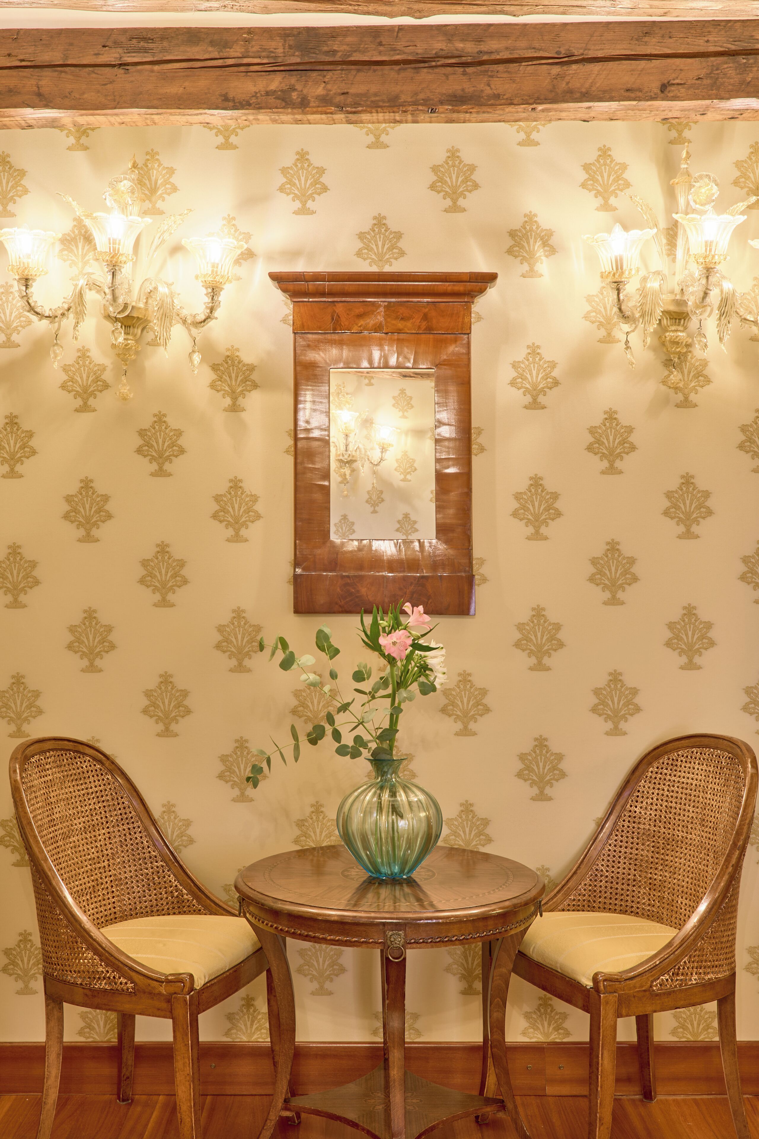 A room with two wicker chairs, a round wooden table with a vase, and wall-mounted lights.
