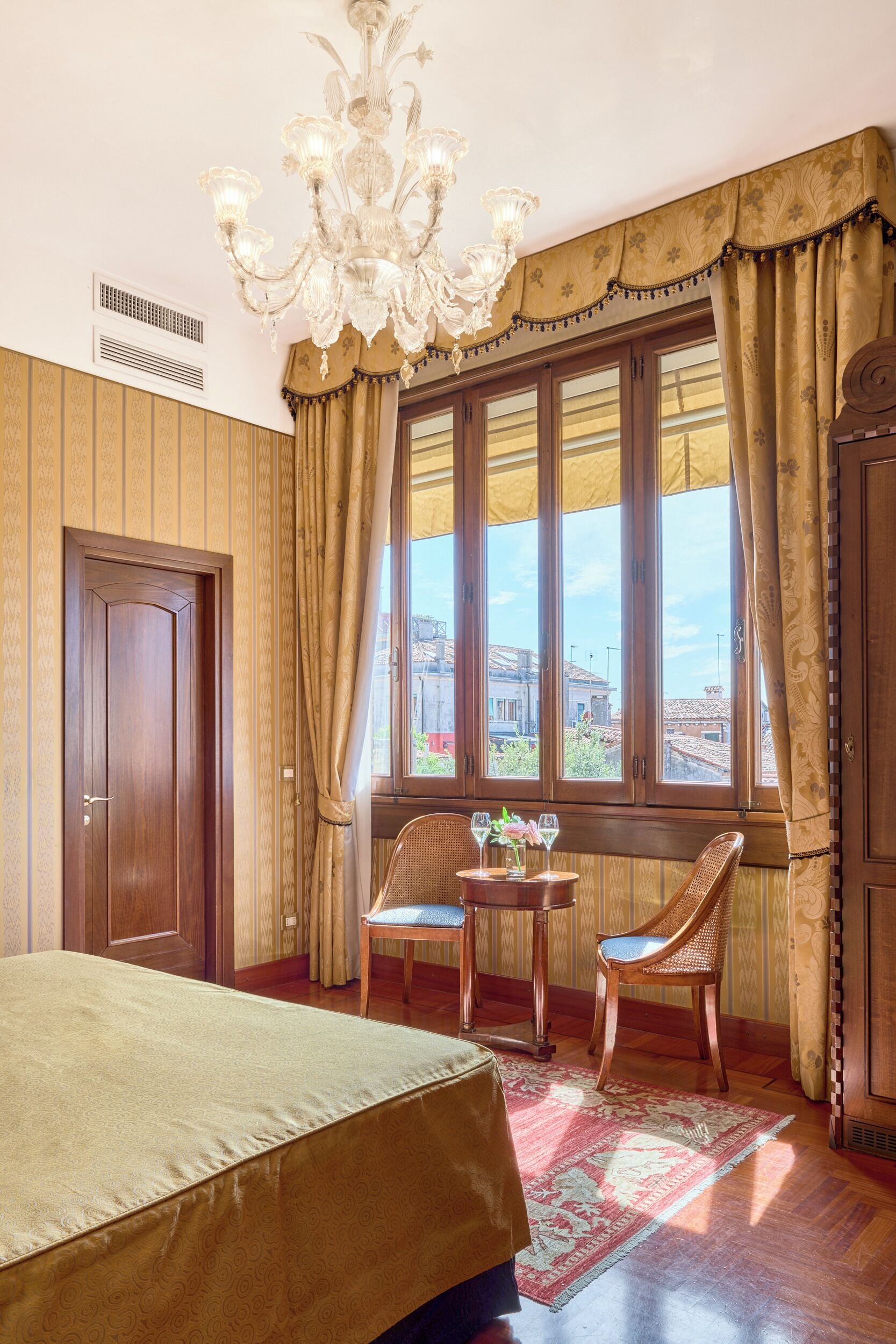 Quarto com cama, duas cadeiras, mesa com copos, lustre e janela com cortinas.