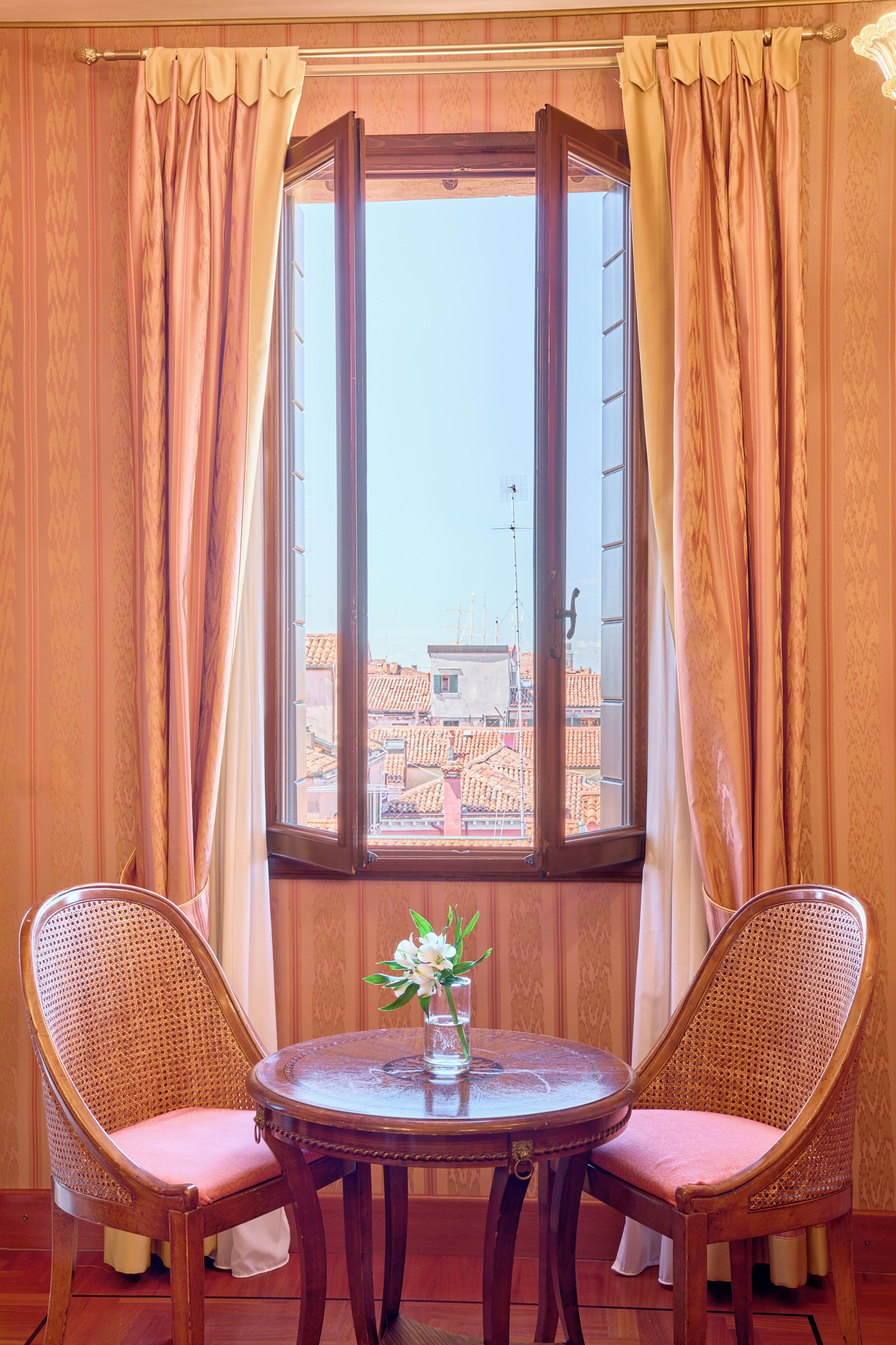 Una habitación con ventana, dos sillas de mimbre, una mesita redonda con un florero y paredes de madera.