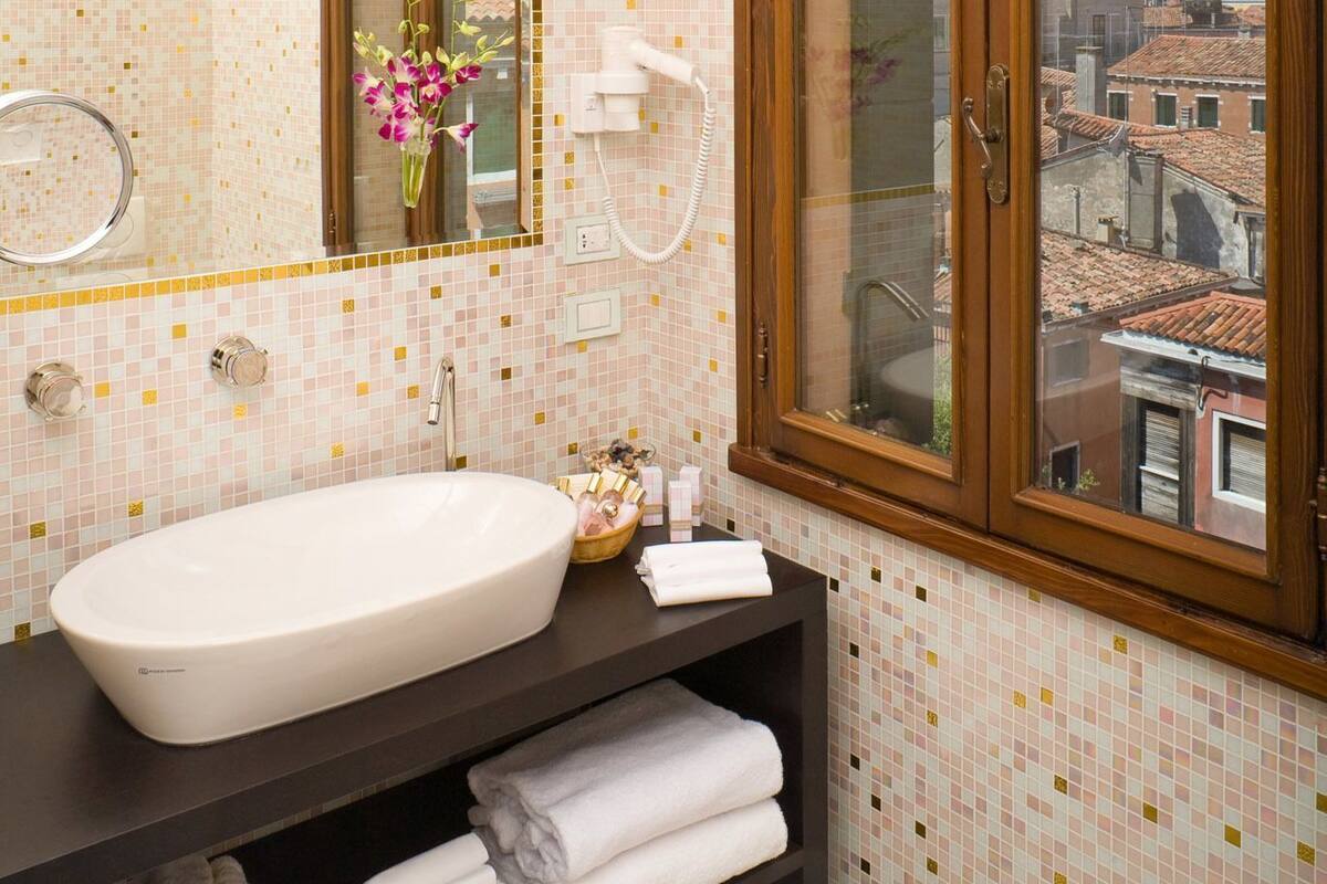 A bathroom with a large mirror, a sink, a window with a view of buildings, and a shelf with towels.