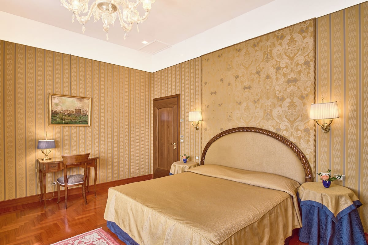 A hotel room with a bed, two bedside tables with lamps, a wooden desk, a chair, a door, and a chandelier.