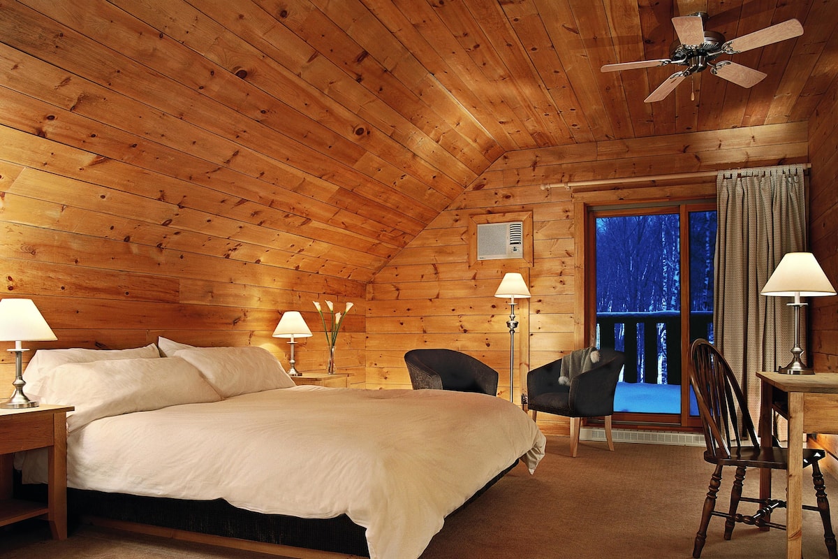 Ein gemütliches Schlafzimmer in einer Holzhütte mit einem großen Bett, zwei Stühlen, einem Tisch und einem Fenster mit Blick auf Bäume.