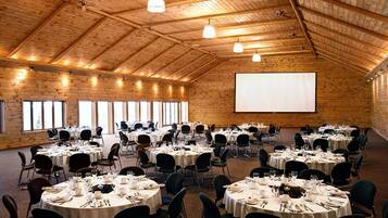 A large hall with multiple round tables set for dining, a projector screen, and wooden walls.