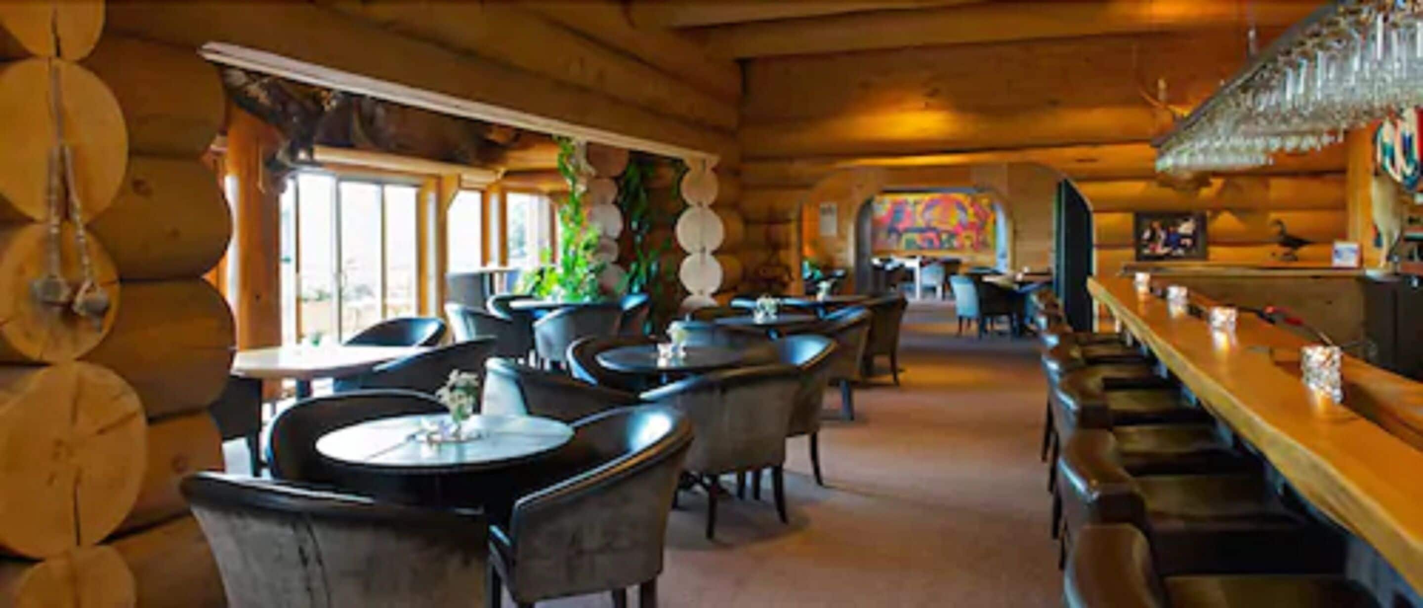 A cozy log cabin-style restaurant with round tables and chairs, a bar area, and a mural on the wall.