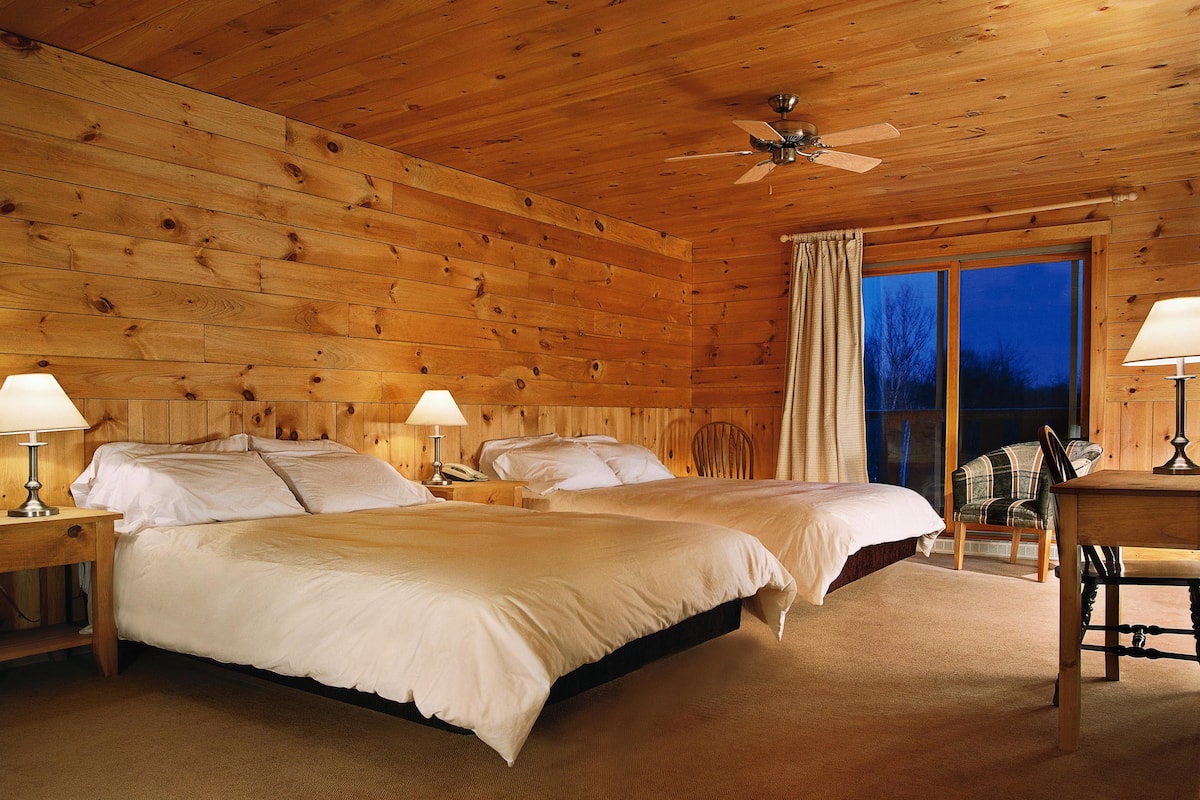 Ein Zimmer in einer Holzhütte mit zwei Betten, einem Deckenventilator und einem großen Fenster.
