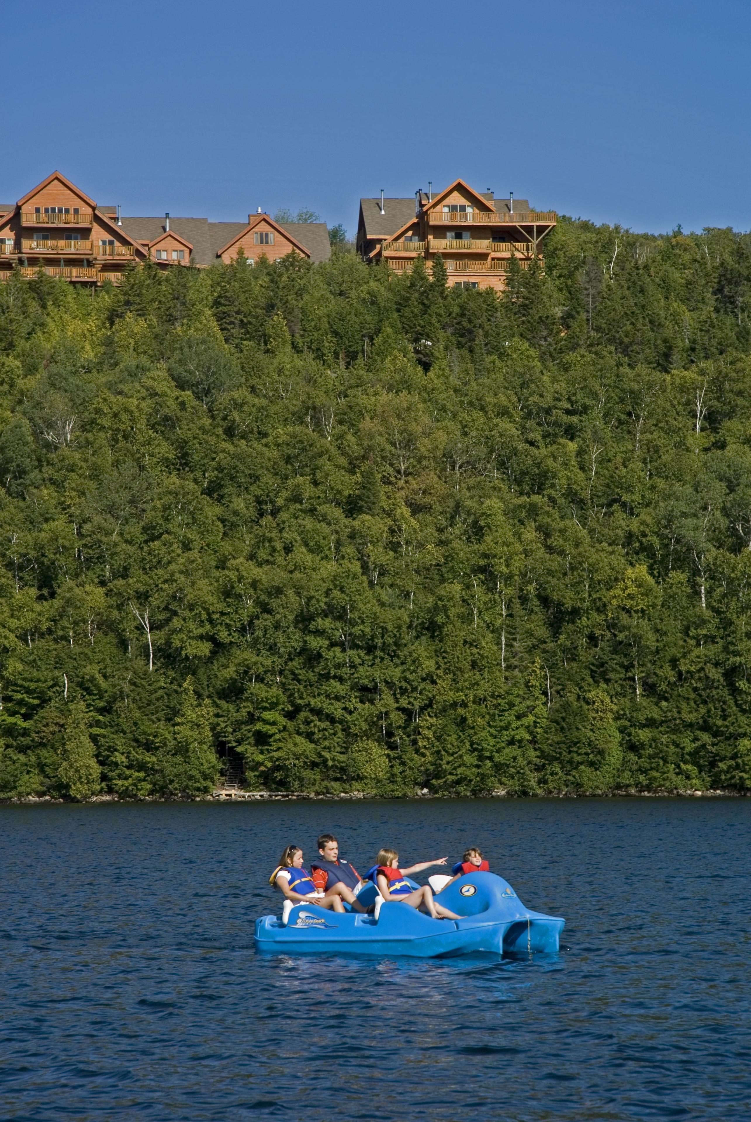 A group of people on a blue boat on a lake with wooden cabins in the background.