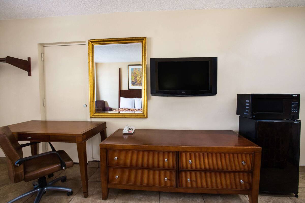 A hotel room with a wooden desk, a chair, a dresser, a television, and a refrigerator.