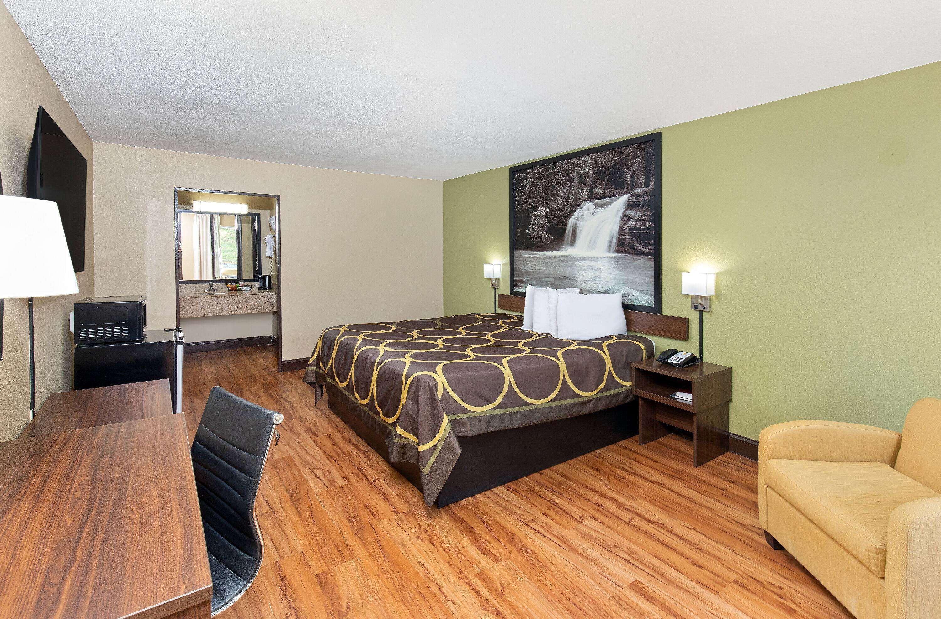 A hotel room with a bed, a desk, a chair, a sofa, a mirror, a nightstand, and a wall-mounted TV.