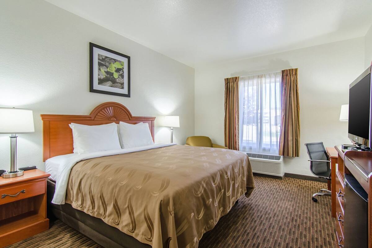 A hotel room with a bed, two bedside tables, a desk with a chair, a television, and a window with curtains.