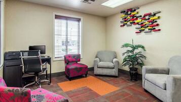 A room with a desk, two armchairs, a small table, a potted plant, and a window with blinds.