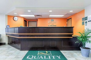 A reception desk with a clock, a sign for Quality Inn by Choice Hotels, and a green carpet with the hotel logo.