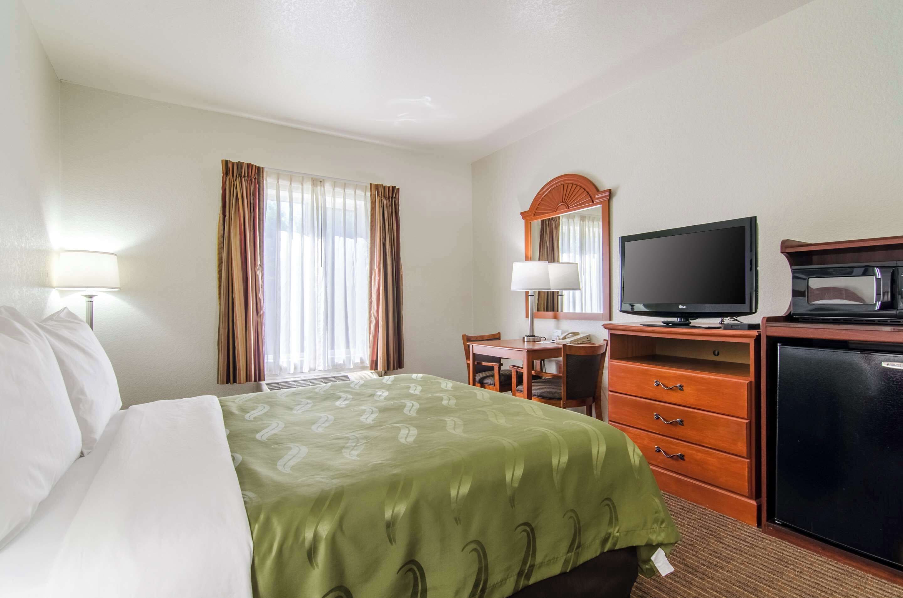 Una habitación de hotel con cama, escritorio, silla, televisión, refrigerador y espejo.