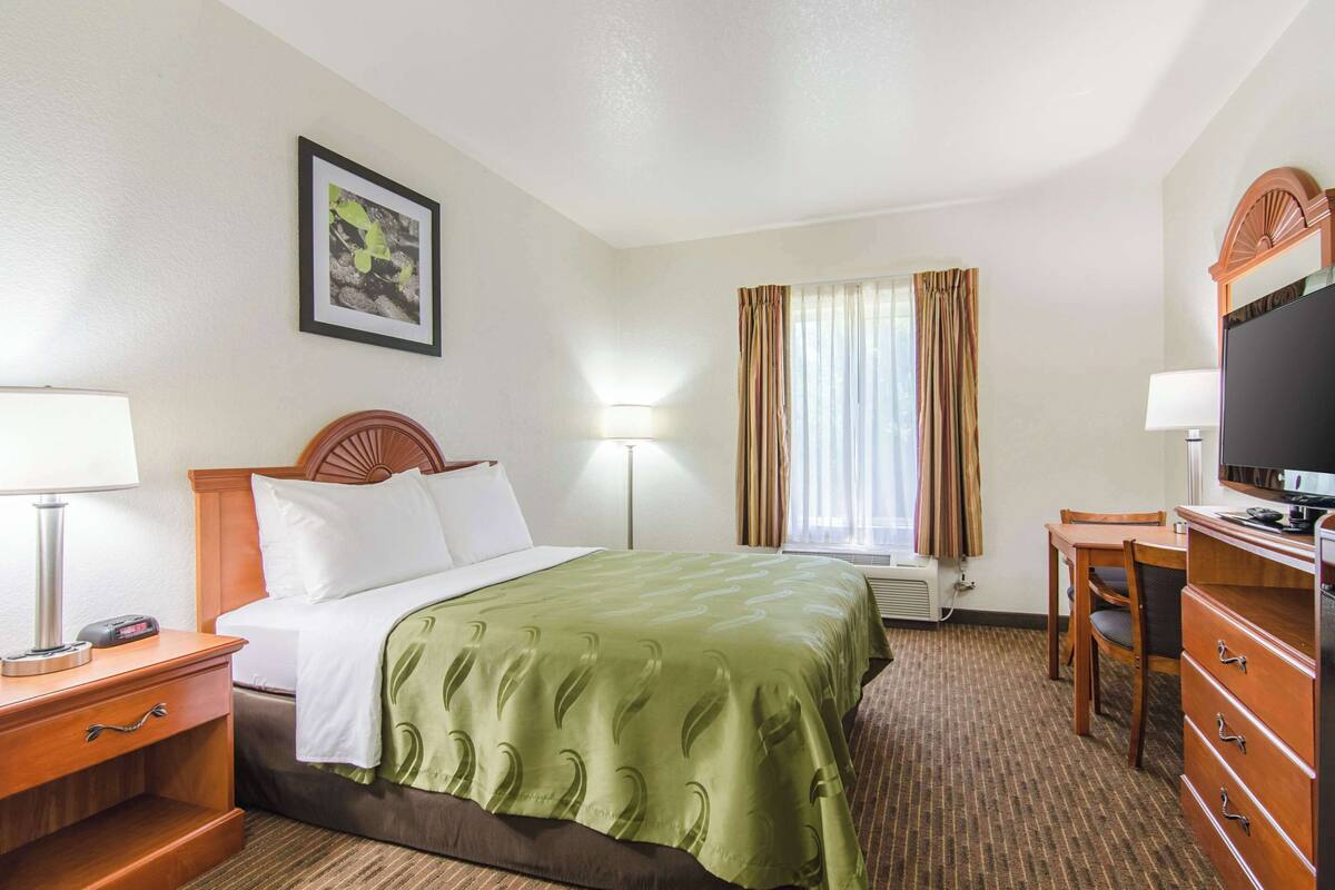 A hotel room with a bed, bedside table, lamp, TV, and a desk with a chair.