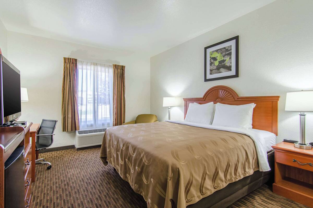 A hotel room with a bed, a desk with a television, a chair, and a window with curtains.