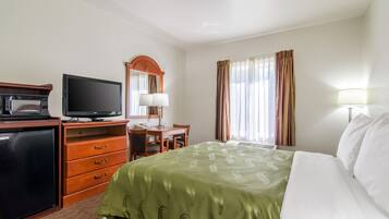 A hotel room with a bed, a desk with a chair, a television, a refrigerator, and a window with curtains.