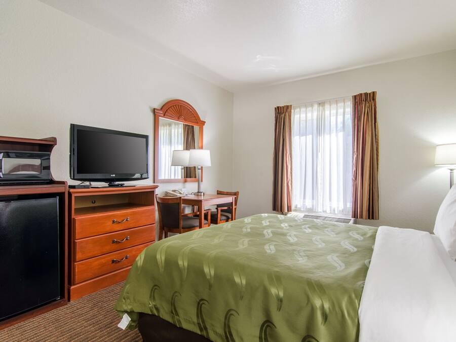 A hotel room with a bed, a desk with a chair, a television, a refrigerator, and a window with curtains.