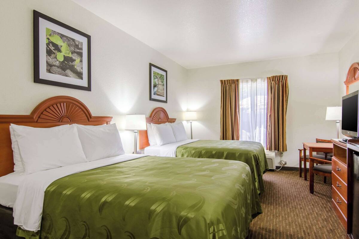 A hotel room with two beds, a desk, a television, and framed pictures on the wall.