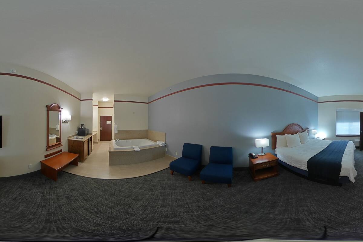 Vista panorâmica de um quarto de hotel com televisão, escrivaninha, área de estar e uma cama com cabeceira.
