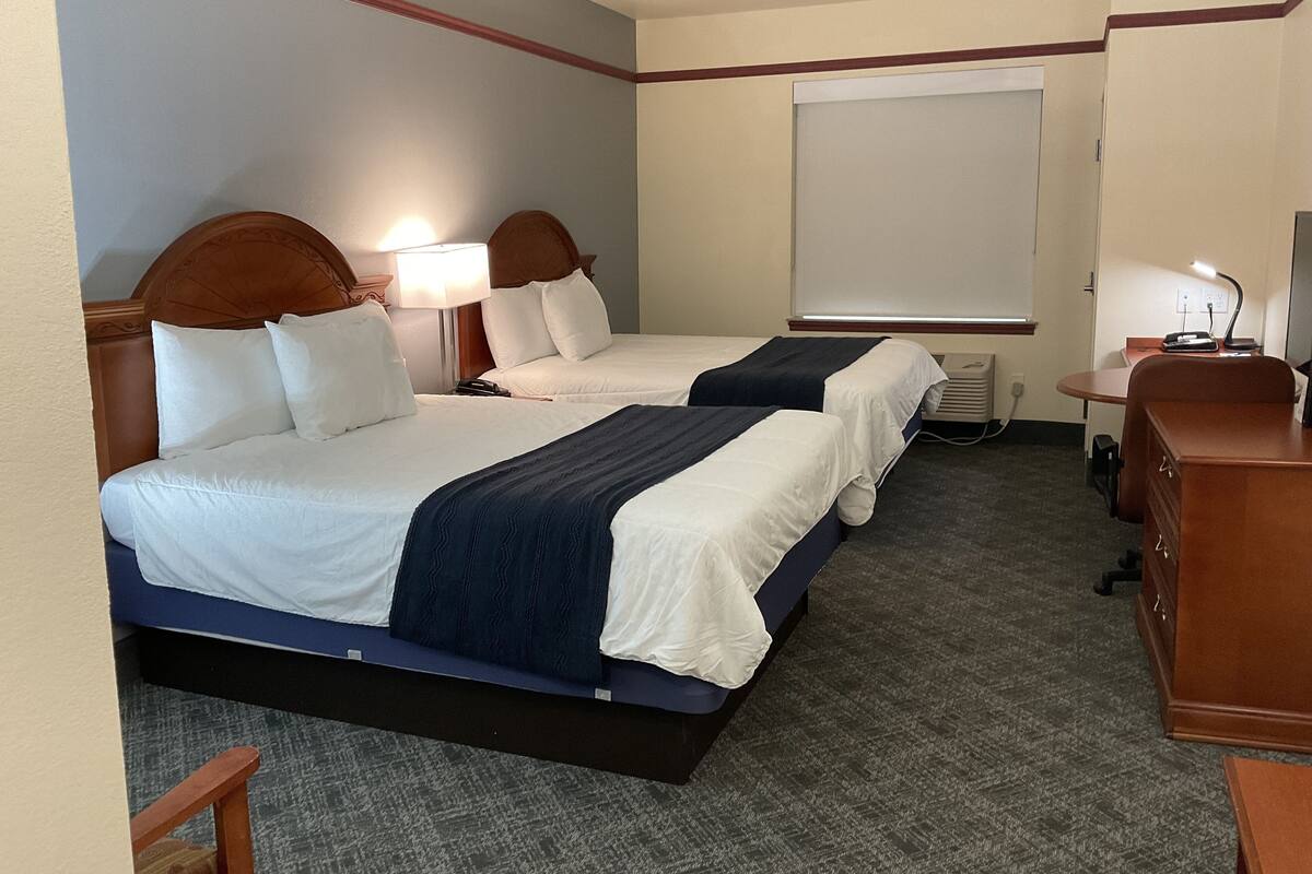 Quarto de hotel com duas camas, uma escrivaninha, uma cadeira, um abajur e uma janela.