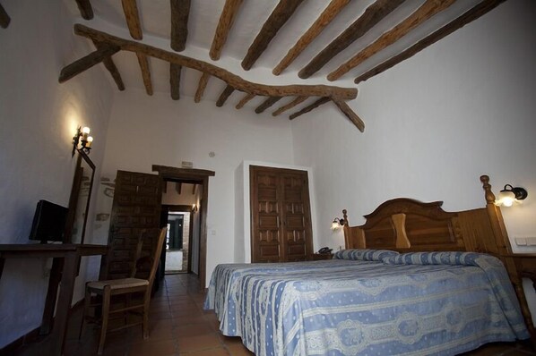 A bedroom with a wooden ceiling, a bed with a patterned bedspread, a desk with a chair, and a television. - Hotel El Horcajo (Montecorto)