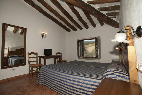 A bedroom with a wooden ceiling, a bed with striped bedding, a desk with a chair, a mirror, and a window with a view of a landscape.