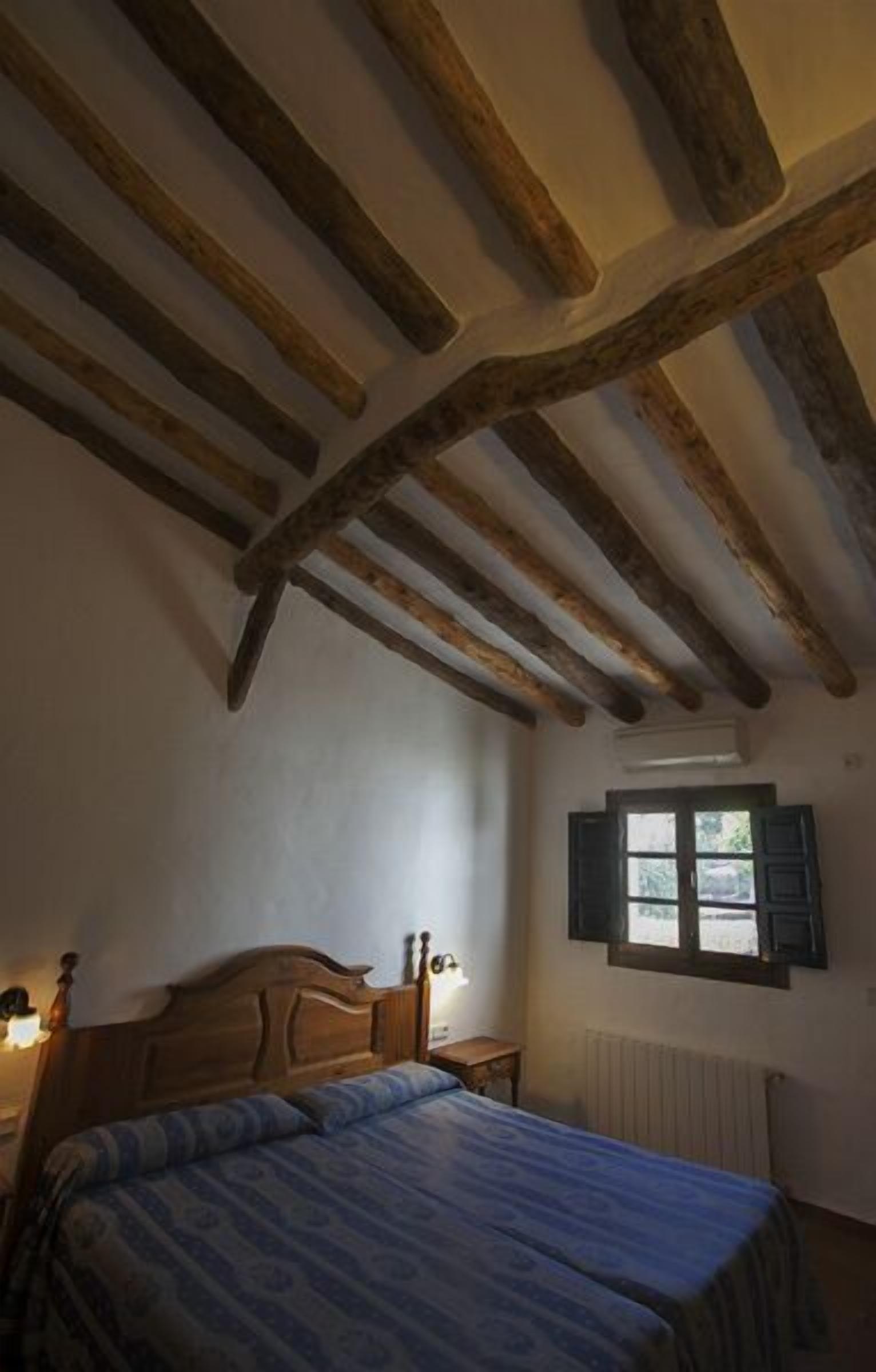 Un dormitorio con techo de madera, una cama con colcha estampada, una mesita de noche con lámpara y una ventana con contraventanas.