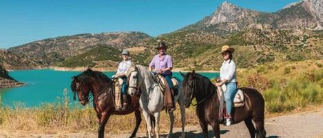 Three people riding horses on a dirt path with a mountainous backdrop and a body of water.