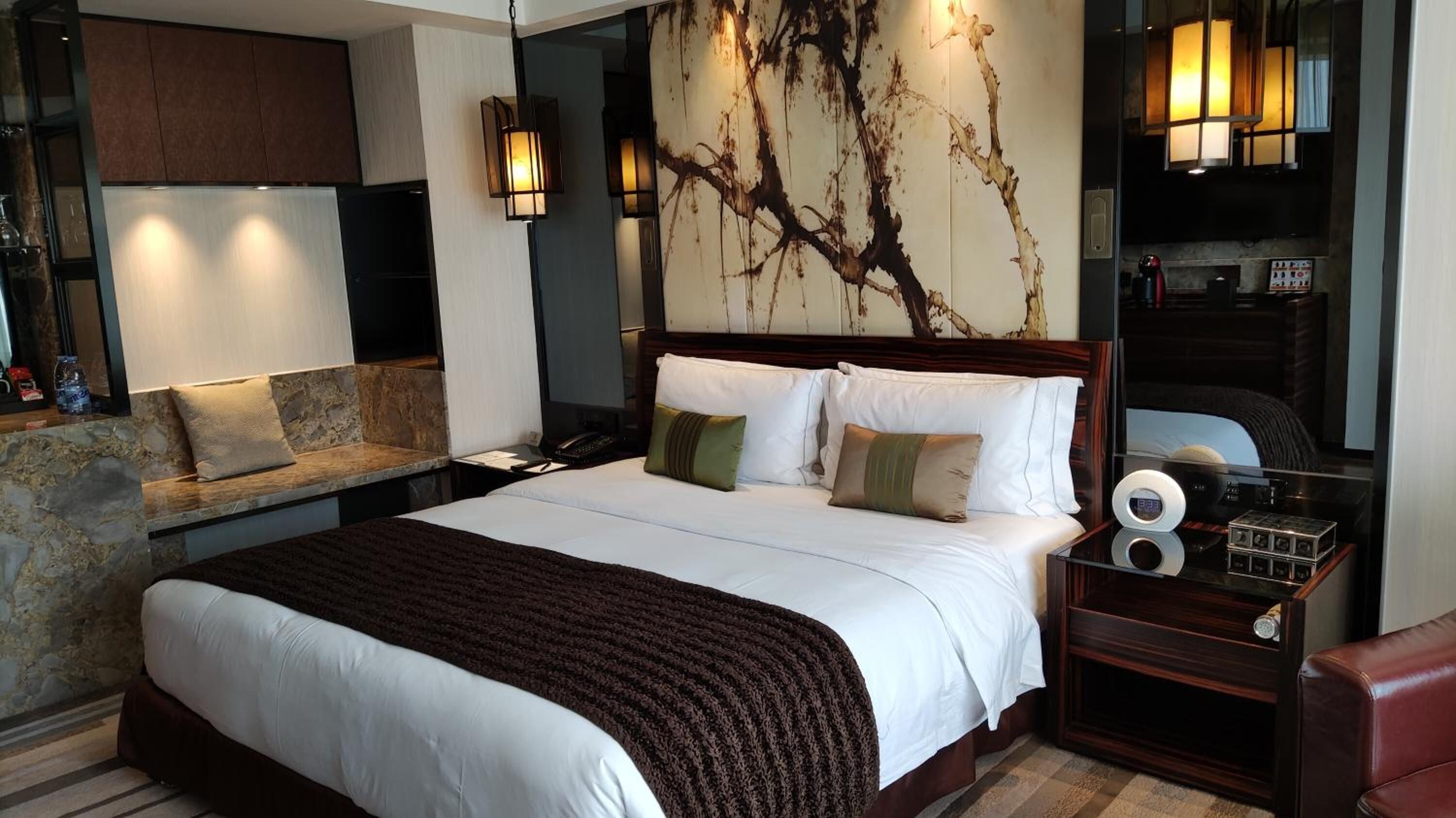 Habitación de hotel con una cama grande, un área de descanso con sofá y sillones, un televisor montado en la pared y una mesita de noche con reloj y altavoz.