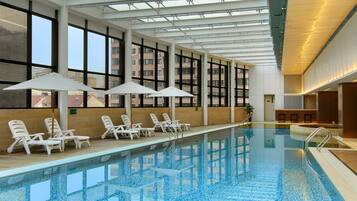 An indoor swimming pool with clear blue water, surrounded by white lounge chairs and umbrellas.