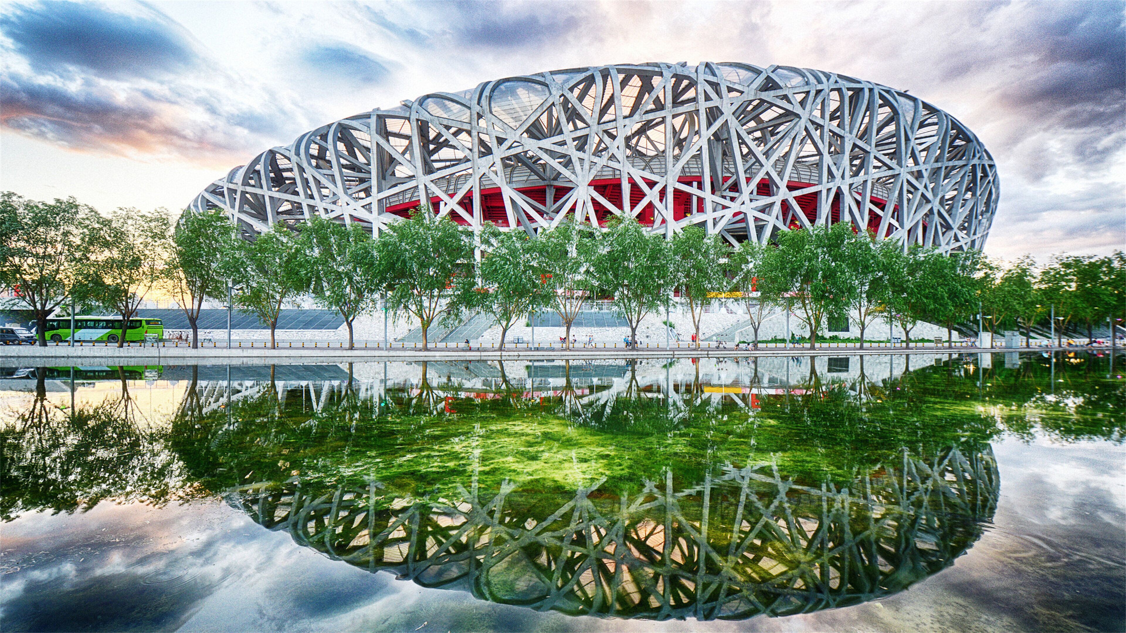 Un estadio grande y circular con una estructura metálica compleja, rodeado de árboles y una masa de agua que refleja la luz.