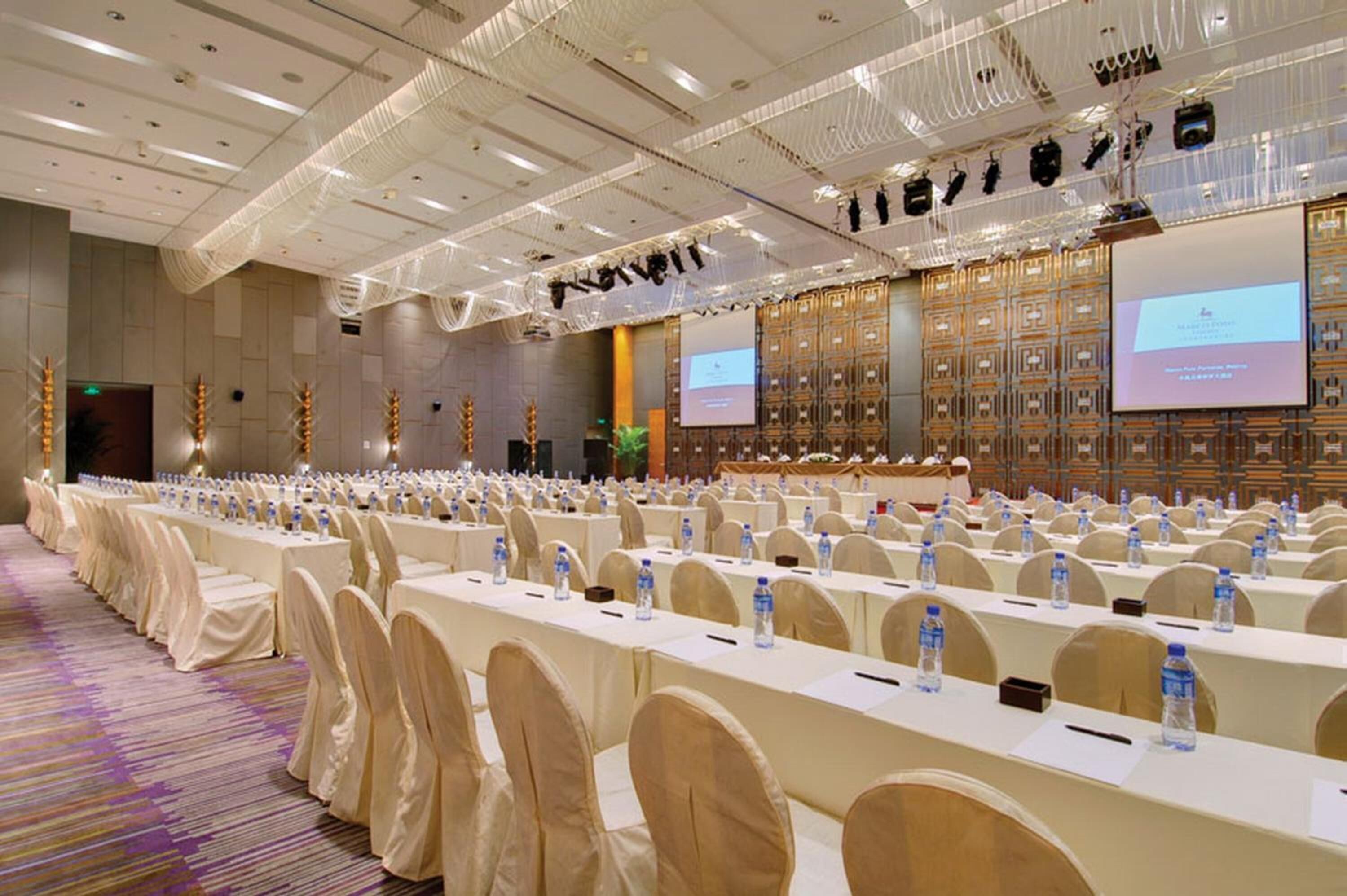 Een ruime congreszaal met rijen tafels en stoelen, voorzien van waterflesjes en plaatskaarten, en een presentatiescherm aan de voorzijde.