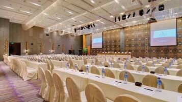 Een ruime congreszaal met rijen tafels en stoelen, voorzien van waterflesjes en plaatskaarten, en een presentatiescherm aan de voorzijde.