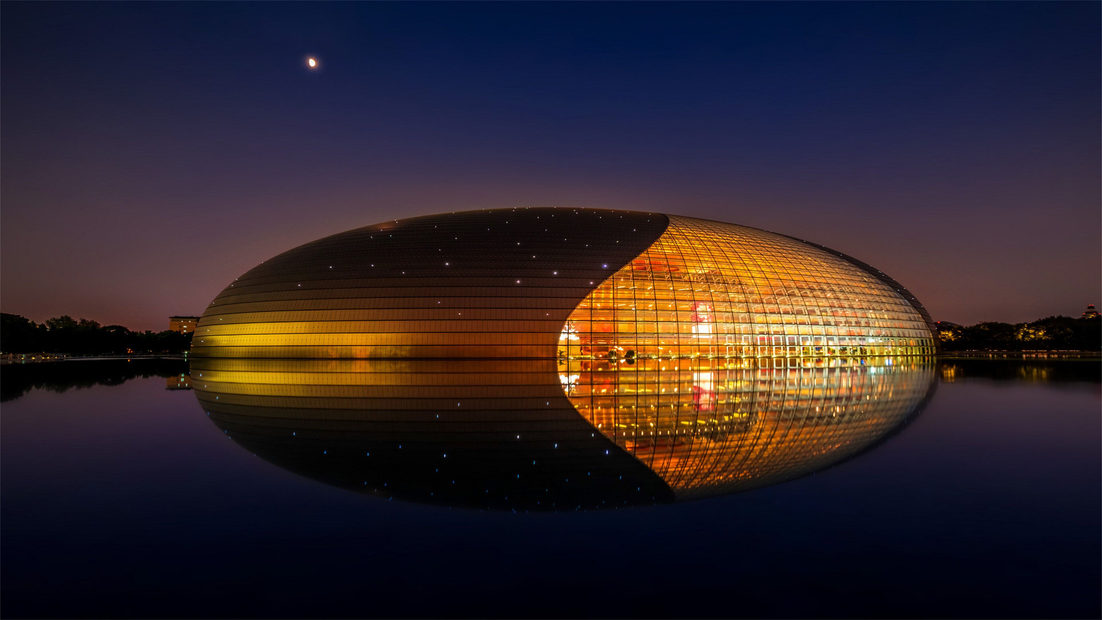 A large, oval-shaped building with a reflective surface, likely a stadium or arena, surrounded by calm water and a night sky.