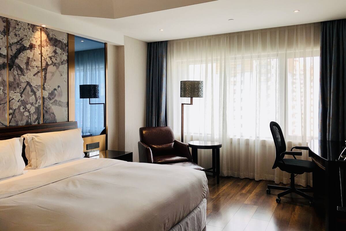A hotel room with a large bed, a desk with a chair, a small table, and a large window with curtains.