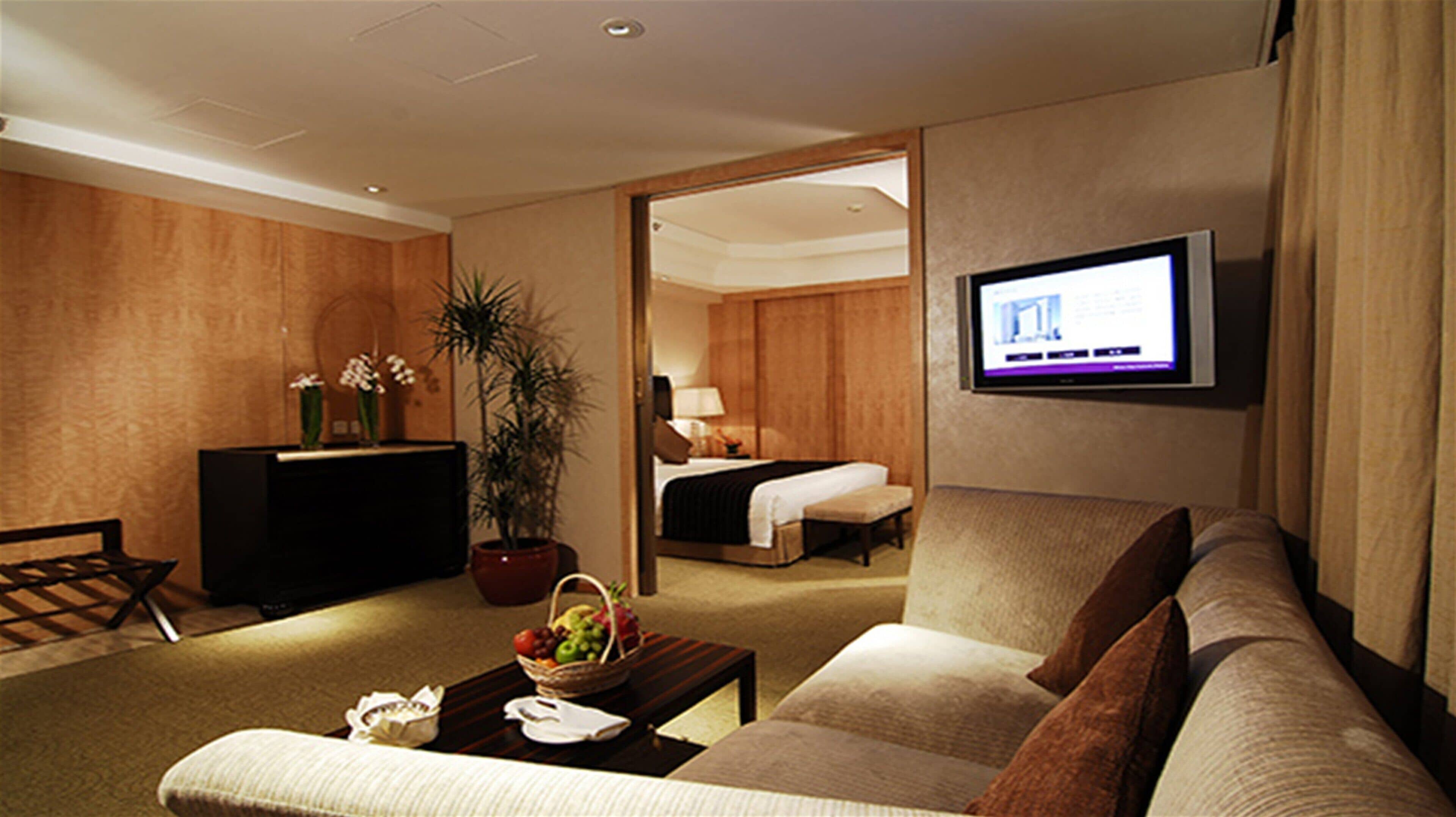 Ein Hotelzimmer mit einer Sitzecke, einem Fernseher, einem Schreibtisch mit Computer sowie einem Bett und einer Zimmerpflanze.