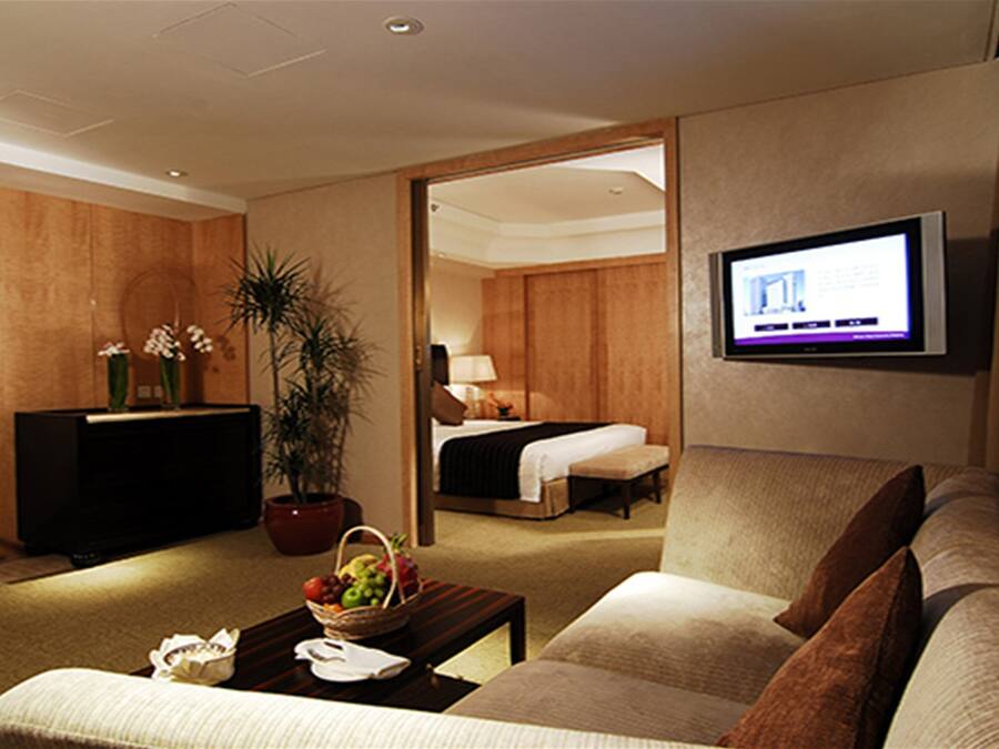 Ein Hotelzimmer mit einer Sitzecke, einem Fernseher, einem Schreibtisch mit Computer sowie einem Bett und einer Zimmerpflanze.