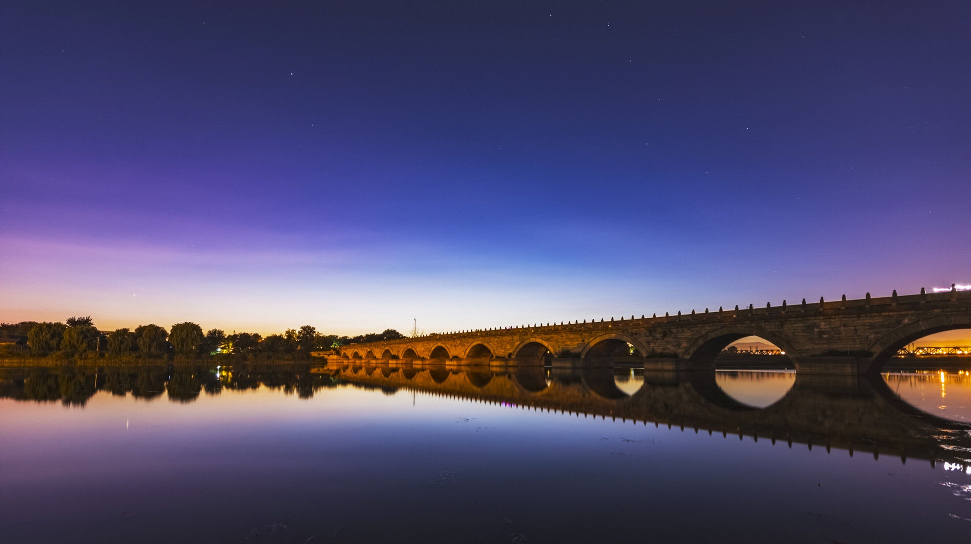 A stone bridge over a calm body of water at twilight, reflecting the bridge and surrounding trees.