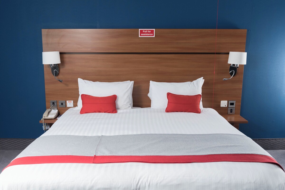 A hotel room with a large bed, two bedside lamps, a phone, and a sign indicating assistance.
