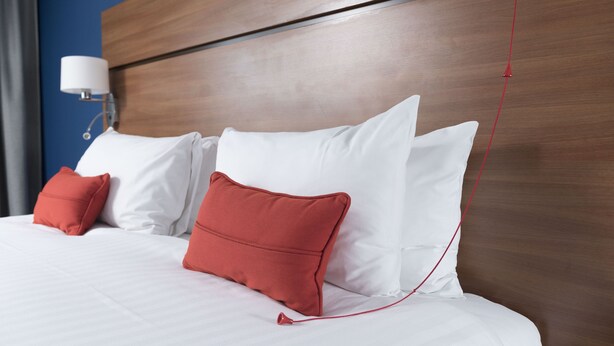 A neatly made bed with white and red pillows, a wooden headboard, and a wall-mounted lamp. - Holiday Inn Express Birmingham Star City by IHG (Birmingham)