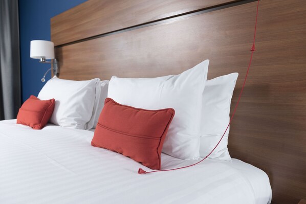 A neatly made bed with white and red pillows, a wooden headboard, and a wall-mounted lamp. - Holiday Inn Express Birmingham Star City by IHG (Birmingham)