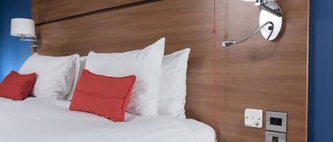 Una habitación de hotel con una cama bien hecha, almohadas rojas y blancas, una lámpara fijada a la pared y una mesita de noche con toma de corriente y panel de control.