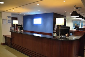 A hotel reception desk with a blue accent wall, a digital display, and a counter with a computer and a telephone. - Holiday Inn Express Birmingham Star City by IHG (Birmingham)