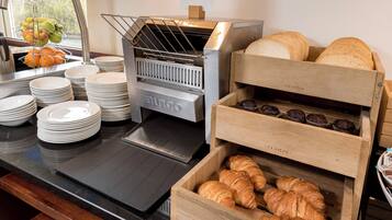 Een ontbijtbuffet met een broodrooster, croissants, muffins en een fruitmand.