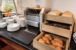 A breakfast buffet with a bread toaster, croissants, muffins, and a fruit basket.