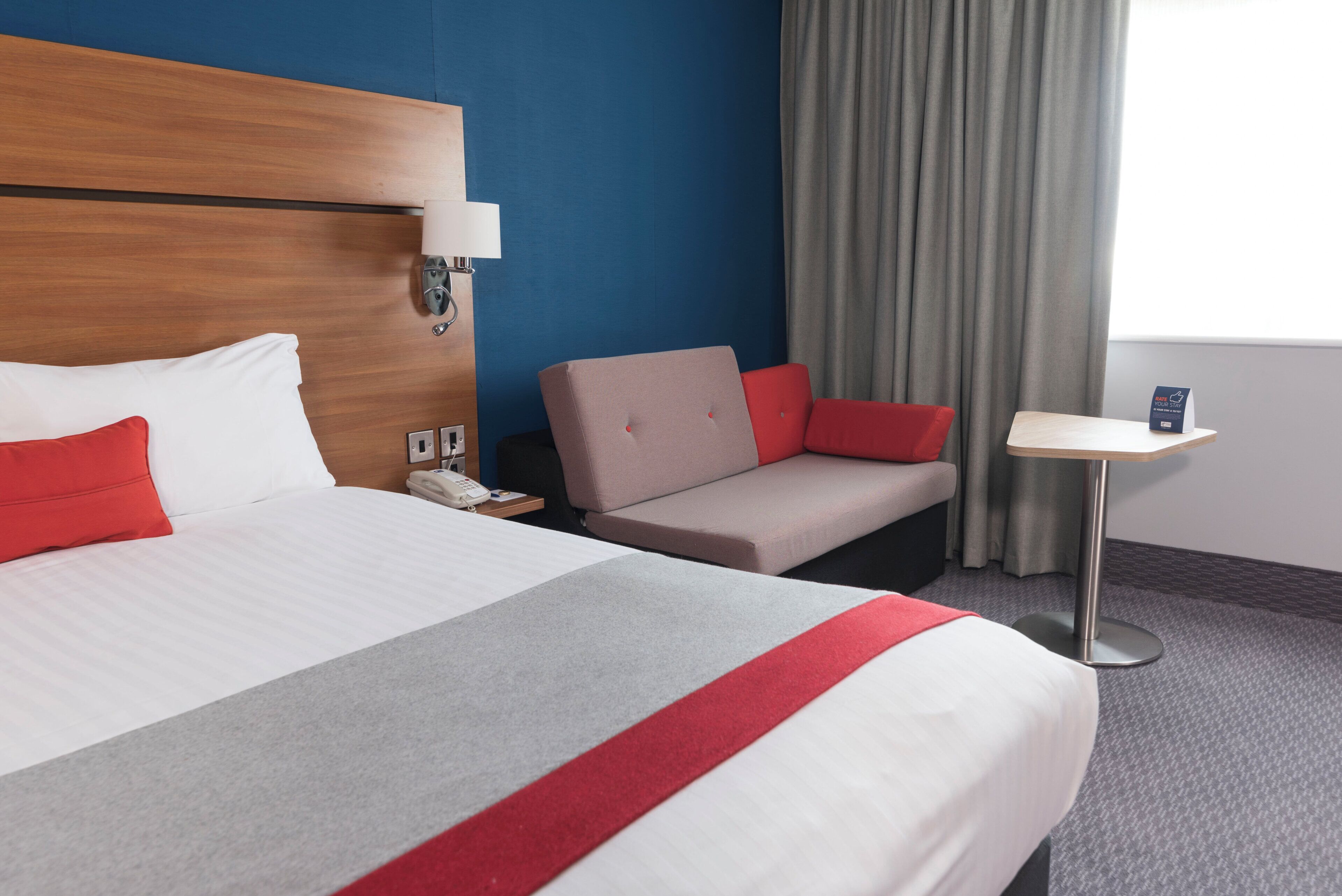 Habitación de hotel con una cama grande, una mesita de noche, una silla, una pequeña mesa, una lámpara, un teléfono y una ventana con cortinas.