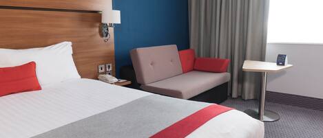 Habitación de hotel con una cama grande, una mesita de noche, una silla, una pequeña mesa, una lámpara, un teléfono y una ventana con cortinas.