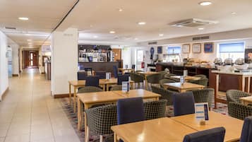 A hotel restaurant with multiple dining tables and chairs, a bar area, and various signs and menus.
