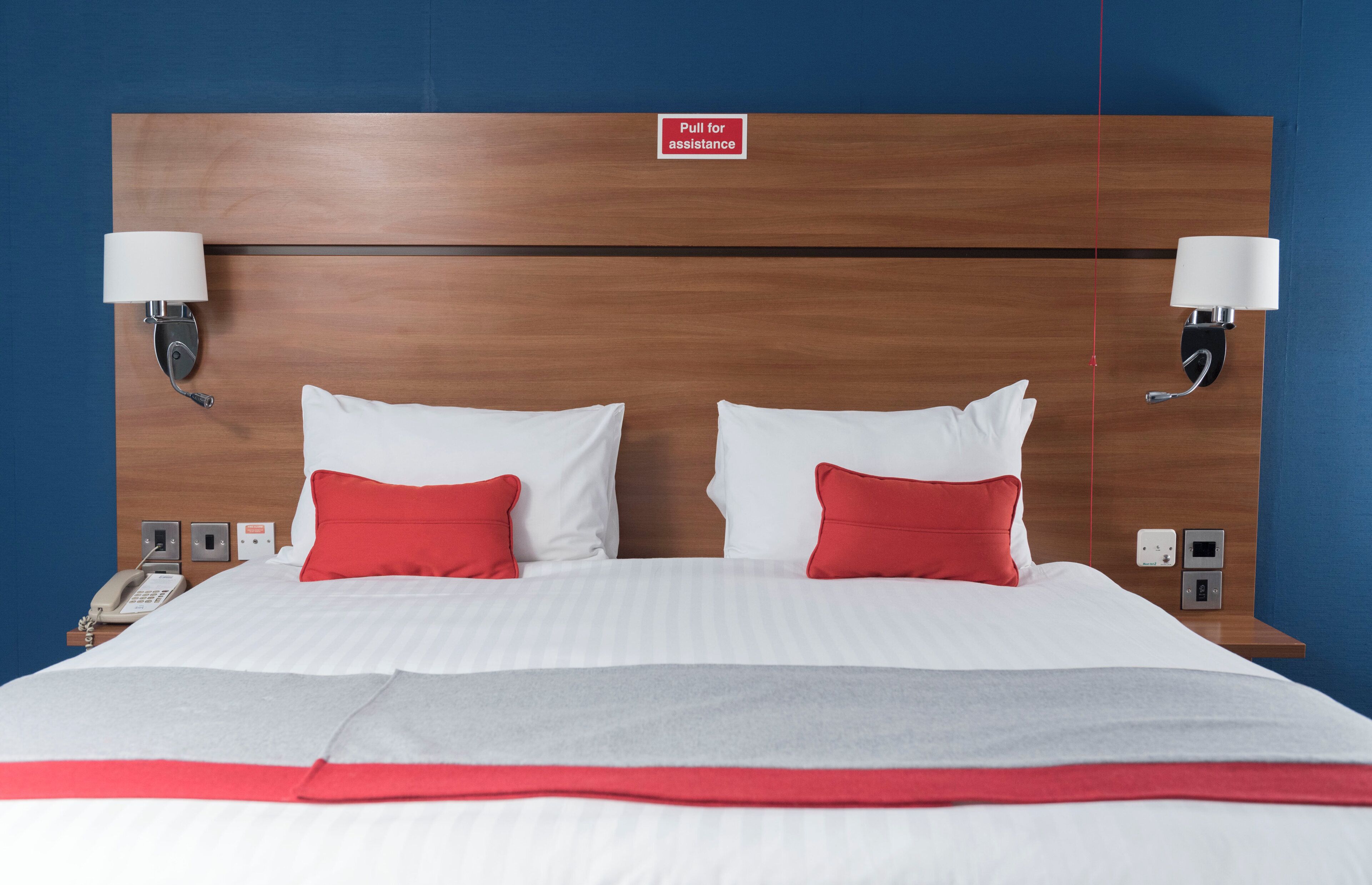 Une chambre d’hôtel avec une tête de lit en bois, deux lampes de chevet, un téléphone et une couvre-lit rouge et blanche.